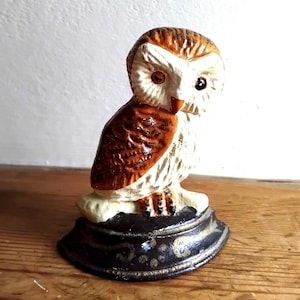 May include: A cast iron owl figurine featuring brown and white painted feathers, a white face with one orange and one black eye, and a small orange beak. The owl is perched on a dark, curved base decorated with subtle gold-toned floral patterns.