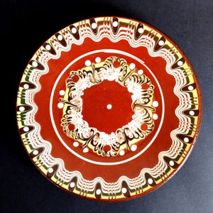 Può includere: Un piatto decorativo in ceramica con una base di colore marrone-rossastro intenso. Il piatto presenta motivi intricati in bianco, marrone e giallo, creando un design visivamente accattivante. Il bordo ha un bordo smerlato e il centro ha un motivo circolare.