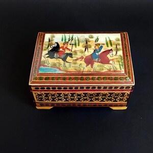 May include: A rectangular wooden box featuring a hand-painted lid depicting two figures riding horses in a landscape. The sides of the box are decorated with intricate geometric marquetry patterns in shades of brown, black, and cream with green accents.