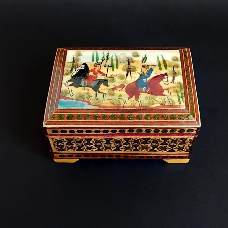 May include: A rectangular wooden box featuring a hand-painted lid depicting two figures riding horses in a landscape. The sides of the box are decorated with intricate geometric marquetry patterns in shades of brown, black, and cream with green accents.