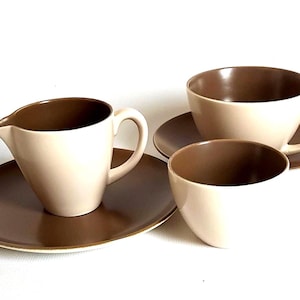 Op de afbeelding: Een set keramisch servies bestaande uit een melkkannetje met handvat en tuit op een schoteltje, een theekopje op een schoteltje en een klein kommetje. Elk stuk heeft een gladde, crèmekleurige buitenkant en een contrasterende donkerbruine binnenkant.