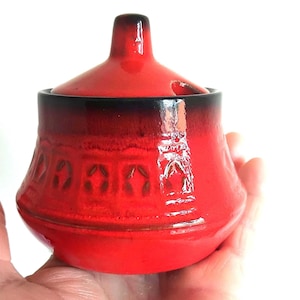 Può includere: Una zuccheriera in ceramica rosso brillante con coperchio abbinato, caratterizzata da un bordo con sfumatura nero scuro e una fascia strutturata con motivi geometrici ripetuti attorno al centro. La ciotola ha una finitura liscia e lucida.