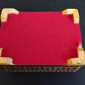May include: A rectangular box featuring a vibrant red felt lining on the top surface, framed by four light-colored wooden corner supports. The sides of the box display a dark, intricate geometric pattern with fine black and white detailing.
