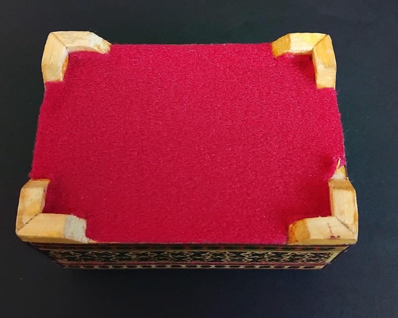 May include: A rectangular box featuring a vibrant red felt lining on the top surface, framed by four light-colored wooden corner supports. The sides of the box display a dark, intricate geometric pattern with fine black and white detailing.