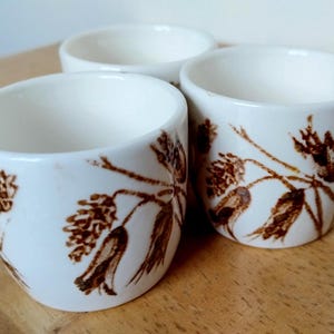 May include: Three white ceramic cups with a brown floral pattern. The cups are cylindrical with a slightly flared rim. The floral design features stylized flowers and leaves. The cups are arranged on a wooden surface.