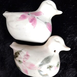 May include: Two white ceramic duck figurines featuring hand-painted floral motifs. The top duck displays pink flower petals, while the bottom duck features a combination of pink petals and green leaves. Both pieces have smooth, rounded bodies.
