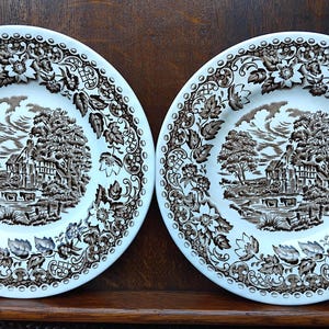May include: Two round ceramic plates featuring a brown and white transferware pattern. The center depicts a rural landscape with a house, trees, and cattle. The wide rim is decorated with a detailed floral and leaf border design.