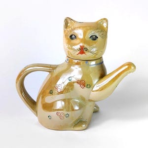May include: A ceramic teapot shaped like a seated cat with a glossy, iridescent beige finish. The cat features painted details including a red nose, dark eyes, and small floral patterns in red, green, and blue. A curved handle and a spout extend from the body.