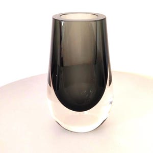 May include: A glass vase featuring a tapered cylindrical shape with a thick, clear base that transitions into a dark, smoky gray upper section. The vessel has a smooth, polished surface and a wide, circular opening at the top, showcasing a clean, modern aesthetic with a distinct contrast between the transparent bottom and the deep gray glass.