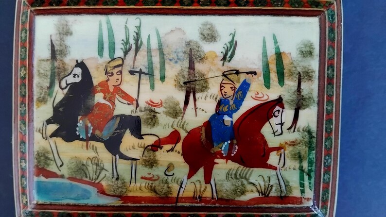 May include: A rectangular decorative box featuring a hand-painted scene of two figures on horseback. One figure wears a red garment on a black horse, while the other wears a blue garment on a red horse. The background depicts trees and a small blue pond.