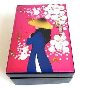 May include: A rectangular black box with a vibrant pink lid featuring a painted illustration of a figure wearing a conical hat and a blue garment. The design includes white cherry blossom flowers and small green leaves against the bright pink background.