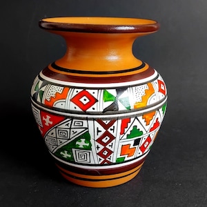 Może przedstawiać: Mały ceramiczny wazon z szeroką szyjką i zaokrąglonym korpusem. Powierzchnia ozdobiona jest ręcznie malowanym geometrycznym wzorem w odcieniach pomarańczu, czerwieni, zieleni, czerni i bieli, z poziomymi pasami wokół górnej i dolnej krawędzi.