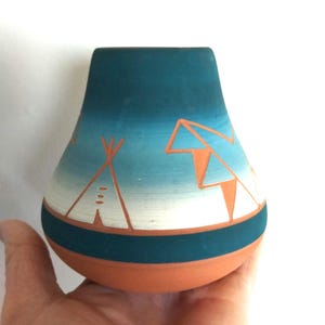 May include: A handmade ceramic vase in teal, blue, and orange. The vase has a gradient design and is decorated with white and orange geometric and teepee-style patterns. The vase is held in a hand.