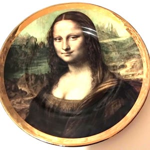 Op de afbeelding: Ronde decoratieve schotel met een reproductie van het schilderij Mona Lisa. De afbeelding van de vrouw staat in het midden van de schotel, met een landschapsachtergrond. De schotel heeft een goudkleurige rand.