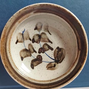 Könnte beinhalten: Eine runde Keramikschale mit gesprenkeltem cremefarbenem Innenraum und braunem Rand. Die Schale hat ein Blumendesign in Braun und Blau. Der Rand hat zwei konzentrische braune Bänder. Die Schale wird wahrscheinlich zum Servieren von Speisen verwendet.