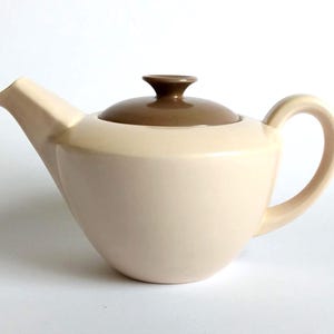 May include: A ceramic teapot with a smooth, matte cream-colored body, a curved handle, and a pointed spout. The teapot features a removable, circular lid in a contrasting solid brown color with a small, central knob for easy handling.