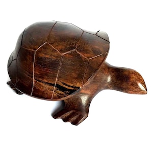 Puede incluir: Una figura de tortuga de madera de color marrón oscuro con un acabado pulido. El caparazón presenta segmentos geométricos tallados en la superficie, mientras que la cabeza y las aletas son lisas y redondeadas. La veta es visible.