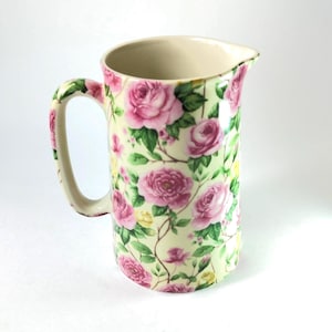 Pode incluir: Jarro de cerâmica com interior e alça creme. O exterior é decorado com um padrão floral de rosas cor-de-rosa, folhas verdes e detalhes amarelos sobre um fundo amarelo pálido. O jarro tem um bico para facilitar o despejo.