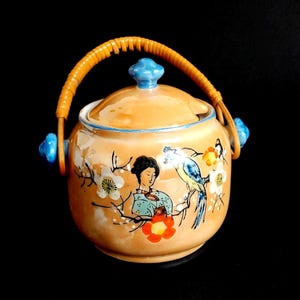 Puede incluir: Un tarro de cerámica vintage con tapa y asa. El tarro es de color melocotón con un diseño pintado de una mujer, un pájaro y flores. El asa es trenzada y la tapa tiene un pomo azul. El tarro está sobre un fondo negro.