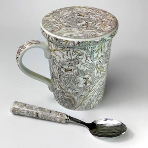 May include: A ceramic tea mug with a matching lid and spoon. The mug, lid, and spoon handle feature a floral pattern in white, gray, and gold. The spoon has a silver-colored bowl.