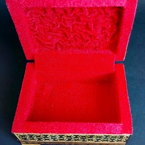 May include: A rectangular wooden box with an open lid, featuring a vibrant red velvet interior lining. The exterior sides and the top edge of the lid are decorated with a detailed, dark-toned geometric inlay pattern in shades of black and gold.