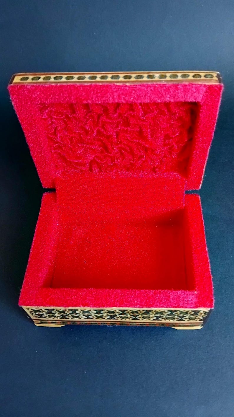 May include: A rectangular wooden box with an open lid, featuring a vibrant red velvet interior lining. The exterior sides and the top edge of the lid are decorated with a detailed, dark-toned geometric inlay pattern in shades of black and gold.