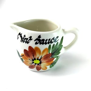Op de afbeelding: Een kleine, witte keramische creamer met een handvat en tuit. De creamer is versierd met een grote, kleurrijke bloem en de woorden "Mint Sauce" in zwart schrift. Groene bladeren accentueren de bloem.