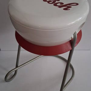 Op de afbeelding: Een witte ronde kruk met een rood Grolsch-logo op het bovenoppervlak. De kruk is voorzien van een rode cirkelvormige steunring onder de zitting en een zilverkleurig metalen frame met gebogen poten die een stabiele basis voor de constructie vormen.