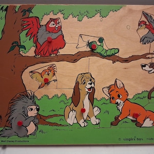 May include: Vintage wooden jigsaw puzzle featuring characters from a Disney movie. The puzzle includes a tree, owl, fox, dog, and other woodland creatures. The puzzle pieces have red knobs for easy handling. The base is green and the puzzle is made in Holland.
