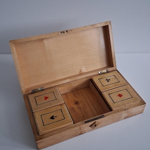 Può includere: Scatola di gioco di carte in legno chiaro, aperta per rivelare quattro piccole scatole di legno, ciascuna con un simbolo di seme: cuori, picche, fiori e quadri. La scatola è in legno chiaro e ha una chiusura in ottone.
