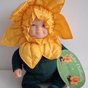 May include: A plush doll in the shape of a daffodil flower, with a yellow petal headpiece and a green body. The doll has a smiling face and a tag that reads "Baby Daffodils by Anne Geddes."