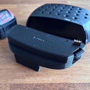 May include: A black 3M Adflo battery pack component with a textured finish and exposed electrical contacts, positioned next to a smaller black and red Milwaukee battery pack. The 3M Adflo unit features a curved, perforated top section.