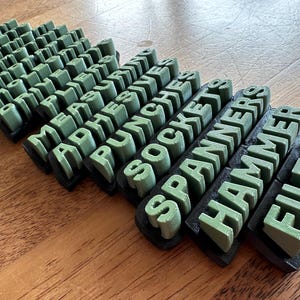Pode incluir: Etiquetas de ferramentas verdes impressas em 3D, incluindo "MEASURE", "ADHESIVE", "PUNCHES", "SOCKETS", "SPANNER", "HAMMER" e "FILES". As etiquetas estão em uma fonte sem serifa e montadas em bases pretas. As ferramentas estão dispostas em uma superfície de madeira.