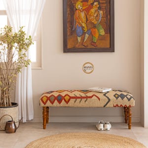 May include: A patterned fabric bench with wooden legs, featuring red, yellow, blue, and beige designs. The bench is in a room with a painting and a round rug. The bench is part of the home decor, with a length of approximately 120 cm.
