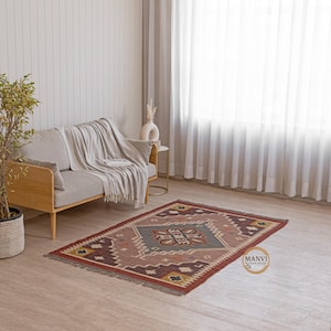 Pode incluir: Um tapete retangular com um padrão geométrico em tons de castanho, cinzento, creme e amarelo. O tapete tem extremidades com franjas e um motivo central em diamante. Um logótipo circular com o texto MANVI HOME DECOR é visível no tapete.