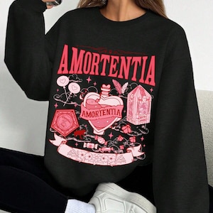 May include: Black sweatshirt with a pink graphic design. The design includes the word "AMORTENTIA" in large letters, a heart-shaped potion bottle, and other decorative elements. The sweatshirt is worn with black pants and white sneakers.