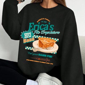 Puede incluir: Sudadera negra con un gráfico de "Erica's Pie Emporium" que muestra una porción de tarta en un plato. El diseño incluye el texto "Served with a side of sass" y "Eat your DAMN PIE!"