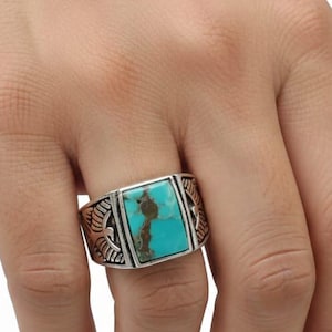 May include: A silver-toned ring with a rectangular turquoise stone. The ring has a detailed design on the sides. The turquoise stone is a vibrant blue-green color. The ring is a statement piece of jewelry.