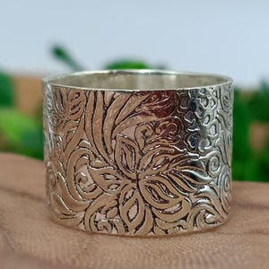 May include: A wide, silver-colored ring with an intricate floral pattern. The ring's surface is covered in a detailed, embossed design of leaves and swirling patterns. The ring is approximately 1 inch wide.