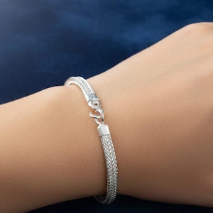 Könnte beinhalten: Ein silbernes Armband mit einem geflochtenen Kettenmuster. Das Armband hat einen sicheren Verschluss und wird an einem Handgelenk vor einem dunkelblauen Hintergrund gezeigt. Es ist ein schlichtes, aber elegantes Schmuckstück.
