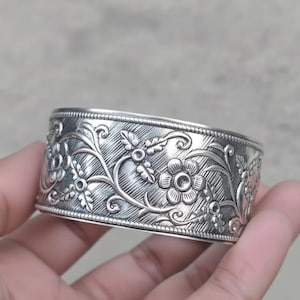May include: A silver cuff bracelet with an intricate floral design. The bracelet features detailed carvings of flowers and leaves, creating a textured surface. The cuff is wide and appears to be made of metal.