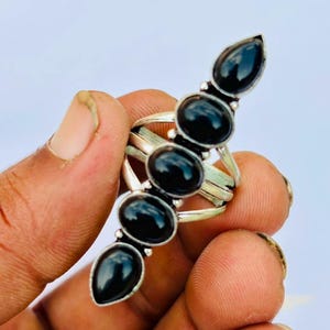 May include: A silver ring featuring five vertically aligned, smooth, black oval-shaped stones. The stones are set in a metallic frame with a split band design, creating a long, slender appearance that extends along the finger when worn.