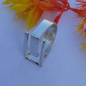 May include: A silver-colored ring with a rectangular top and a U-shaped design. The ring is set against a soft, light background with yellow and orange floral accents.