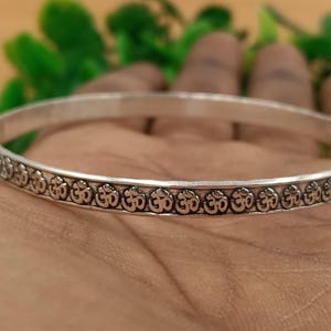 May include: A silver-colored bangle bracelet with an engraved pattern of the Om symbol. The bracelet is round and appears to be made of metal. The Om symbol is repeated around the entire bracelet.