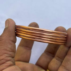 以下が含まれることがあります： 銅色のバングルが積み重なっています。ブレスレットは丸く、無垢の金属でできているようです。バングルは、白い背景を背景に、人の手で持たれています。