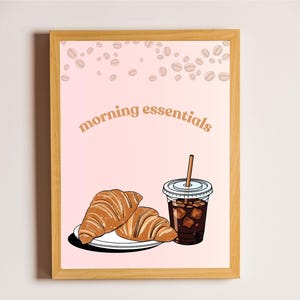 May include: A framed print with the words "morning essentials" in a retro font. The image features two croissants on a plate and a cup of iced coffee. Coffee beans are scattered in the background.