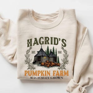 May include: A cream-colored crewneck sweatshirt featuring a graphic print of a stone hut surrounded by pine trees and pumpkins. The text reads HAGRID'S EST. 1981 PUMPKIN FARM MAGICALLY GROWN in green and orange lettering with a decorative leaf border.