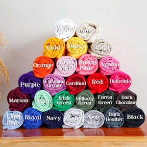 May include: A pyramid of rolled-up t-shirts in various colors, including white, gold, orange, purple, maroon, light blue, royal blue, navy, ash, and sport grey. Each shirt is labeled with its color name, such as "Daisy," "Safety Pink," and "Dark Chocolate."