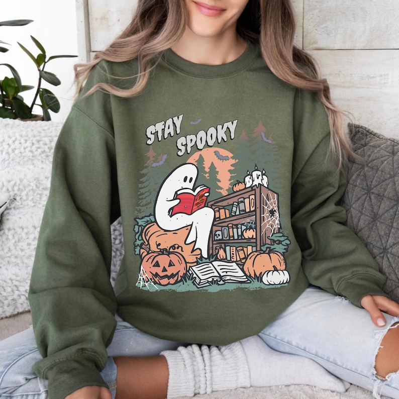 May include: An olive green sweatshirt with a Halloween graphic. The design shows a white ghost reading a red book, surrounded by pumpkins, a bookshelf, and the text "Stay Spooky". The background includes a forest and a full moon.