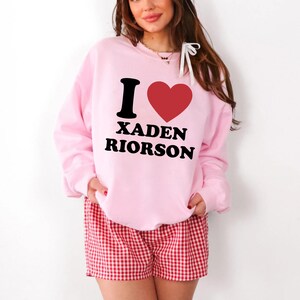 May include: A light pink sweatshirt with the text "I ❤️ XADEN RIORSON" in black and red. The person is wearing red and white checkered shorts. The sweatshirt is a crew neck style.